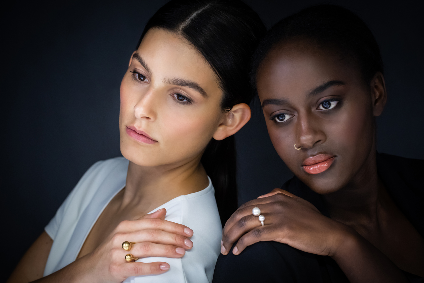 Zwei Frauen mit dem Rücken zueinander und das Gesicht nach vorne gewandt, haben jeweils eine Hand auf ihrer Schulter liegen. An dieser Hand tragen sie einen Zwischenfingerring. Die Ringe sind nach oben hin offen, beschreiben ein U, auf dessen Enden eine Kugel sitzt. Diese liegt dann zwischen den Fingern.