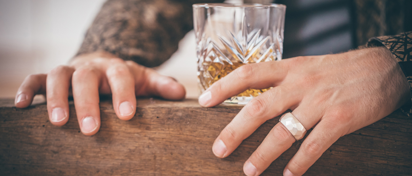 Zwei Männerhände liegen auf einem Holzbalken, in der Mitte ein Whisky Glas. Die lange Hand trägt am Ringfinger einen dicken Silberring. Die Oberfläche ist übersät mit ungleichmäßigen Facetten, die ein interessantes Licht- und Schattenspiel erzeugen.