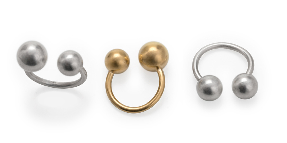 Three rings, each with two balls, lying in a row from a different perspective. The ring band is a U on the ends of which two balls of different sizes face each other. Two rings are silver, the middle one is gold-plated.