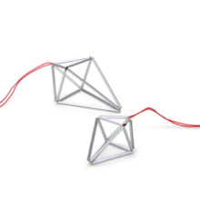 Anhänger Prisma klein und mittel, silberfarben Zwei verschieden große, silberfarbene Anhänger an roter Seidenschnur. Der Anhänger prisma beschreibt einen ungleichseitigen Doppeltetraeder, der an Kristall erinnert. Er ist aus eloxierten Aluminiumröhrchen, die unsichtbar auf Nylon aufgezogen die Seitenkanten des Polyeders bilden.
