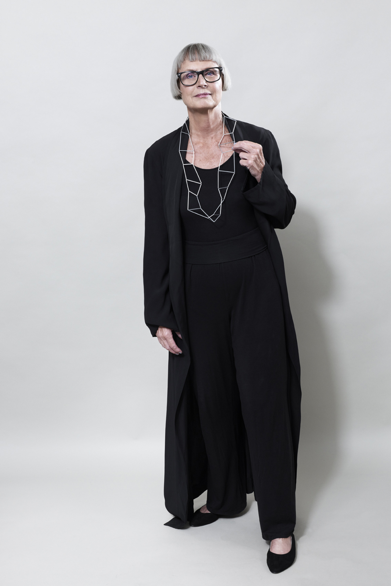 Frau mit lässig elegantem Look in schwarz, trägt einen Halsschmuck, welcher an eine unregelmäßige Leiter erinnert. Entgegen des ersten Eindrucks, scheint die Kette sehr flexibel.