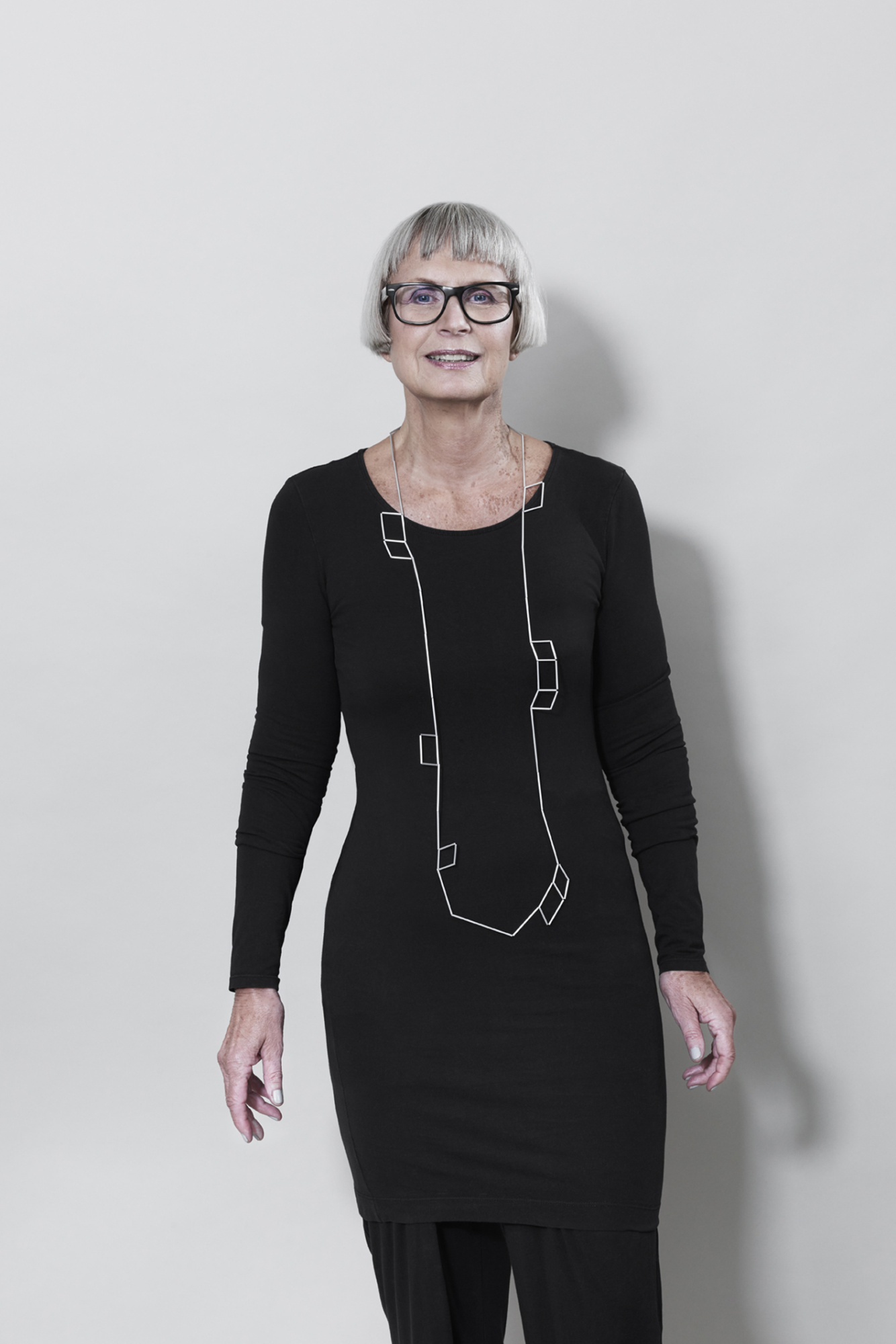 Frau mit kurzem, grauem Haar und einer schwarzen Brille trägt ein schwarzes Kleid über einer schwarzen Hose. Auf dem Kleid trägt sie eine lange Kette bis zum Bauch, welche aus dünnen Aluminiumröhrchen auf Nylon aufgezogen ist. Ähnlich einer Wäscheleine, tanzen in unterschiedlichen Abständen kleine Rechtecke an der Linie entlang. Einzeln, oder auch zwei oder drei nebeneinander.