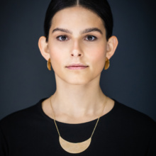 Anhaenger-und-Ohrstecker-moon Frau mit dunklem Haar, streng nach hinten gebunden in frontal Ansicht. Um den Hals trägt sie ein feines Kettchen mit einem großen Anhänger in From einer liegenden Mondsichel. Om Ohr Stecker in Form von Halbkreisen. Sie werden erst aus der Seitenansicht richtig sichtbar. Alles Stücke sind aus vergoldetem Silber gearbeitet.