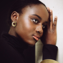 bangle-earrings-courbe-golden Dark-skinned woman leaning against wall. It has an arched gold disc with a circular opening as a stud. The arm is adorned with a gold-colored bangle, also in the form of a curved circular disc.