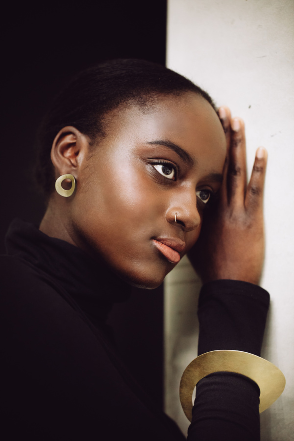 bangle-earrings-courbe-golden Dark-skinned woman leaning against wall. It has an arched gold disc with a circular opening as a stud. The arm is adorned with a gold-colored bangle, also in the form of a curved circular disc.