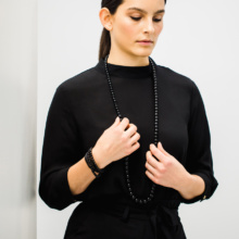 Woman with dark hair and black blouse wears a long chain of different sized onyx balls around her neck. With both hands she almost chained to the chain. On the right wrist she wears another onyx chain wrapped several times.