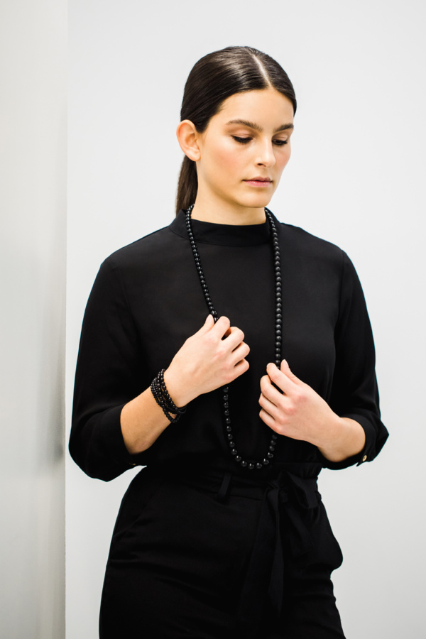 Woman with dark hair and black blouse wears a long chain of different sized onyx balls around her neck. With both hands she almost chained to the chain. On the right wrist she wears another onyx chain wrapped several times.