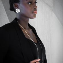 Frau im Profil vor Betonwand. Um den Hals bis auf die Brust trägt sie eine lange, dünne Röhrchenkette. Eine Hälfte der Kette ist geschwärzt, die andere silbern belassen. Im sichtbaren Ohr trägt sie Ohrschmuck in Form einer großen Silberscheibe. Diese ist mit schwarzen Linien aus Faden bespannt.