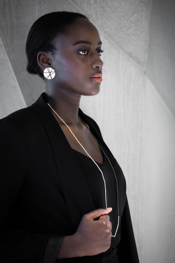 Frau im Profil vor Betonwand. Um den Hals bis auf die Brust trägt sie eine lange, dünne Röhrchenkette. Eine Hälfte der Kette ist geschwärzt, die andere silbern belassen. Im sichtbaren Ohr trägt sie Ohrschmuck in Form einer großen Silberscheibe. Diese ist mit schwarzen Linien aus Faden bespannt.