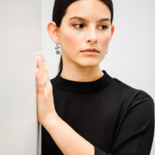 A woman with a black blouse and dark hair is standing on a ledge with her left hand on the wall. The right ear is visible, there she wears earrings. Similar to a panicle, circular plates are arranged on a web.