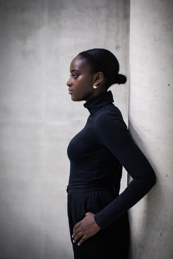 Ohrstecker-GAP-golden Frau lehnt mit dem Rücken gegen eine Säule. Sie trägt goldfarbenen Ohrschmuck in Form von Kreisscheiben. Diese sind bis zur Mitte eingeschnitten, so dass durch auseinander biegen eine räumliche Wirkung entsteht. Eine Spitze sitzt auf dem Ohrläppchen, die andere ihr gegenüber ragt dahinter.