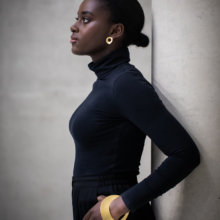 ohrstecker-armreif-courbe-golden Dunkelhäutige Frau mit schwarzem Rollkragenpullover im Profil. Am Ohr ein goldfarbener Ohrschmuck in Form einer gewölbten Scheibe mit einem kreisrunden Durchbruch, allerdings außermittig. Am Handgelenk eine große, ebenfalls vergoldete und in der Mittelachse gewölbte Scheibe mit Kreisdurchbruch als Öffnung für den Arm.