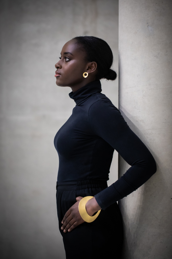 ohrstecker-armreif-courbe-golden Dunkelhäutige Frau mit schwarzem Rollkragenpullover im Profil. Am Ohr ein goldfarbener Ohrschmuck in Form einer gewölbten Scheibe mit einem kreisrunden Durchbruch, allerdings außermittig. Am Handgelenk eine große, ebenfalls vergoldete und in der Mittelachse gewölbte Scheibe mit Kreisdurchbruch als Öffnung für den Arm.