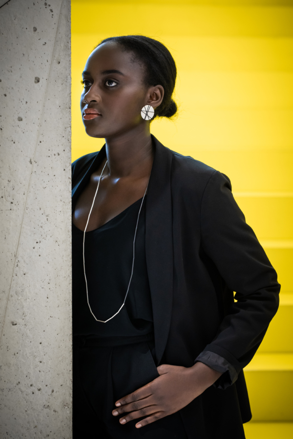 Dunkelhäutige Frau lehnt mit dem halben Oberkörper an Betonsäule. Im Hintergrund eine gelbe Treppe. Sie trägt auffälligen Ohrschmuck. Große Scheiben aus Silber, welche mit einem grafischen Muster aus schwarzem Seidenfaden bespannt sind. Um den Hals über ihre schwarze Kleidung eine Kette aus dünnen Röhrchen. Die eine Hälfte der Röhrchen ist silbern, die andere Hälfte geschwärzt.