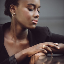 Dark-skinned woman in a black jacket is sitting, hands up and one over the other. On the ring finger of her right hand she wears three golden, delicate rings with a bead look. Her head is turned to the side and only the right ear is visible. There she wears a small, gold ear stud, also a lot of small balls closed in a circle.
