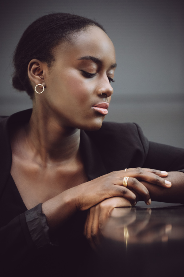 Dunkelhäutige Frau mit schwarzem Jacket sitzt, die Hände aufgestützt und übereinander gelegt. Am Ringfinger der rechten Hand trägt sie drei goldene zarte Ringe in Kügelchenoptik. Ihr Kopf ist zur Seite geneigt und nur das rechte Ohr sichtbar. Dort trägt sie einen kleinen, goldenen Ohrstecker, ebenfalls lauter kleine Kügelchen zum Kreis geschlossen.