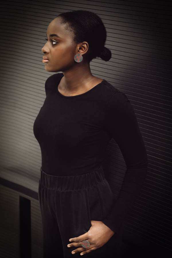 Ohrstecker-und-Ring-big-disk Frau lehnt mit dem Rücken an einem Geländer vor einer dunklen Wand. Den Daumen ihrer linke und sichtbaren Hand hat sie so in die Hosentasche gesteckt, dass der Rest der Hand sichtbar ist. Auf dem Ringfinger trägt sie einen großen Ring. Im linken Ohr einen auffälligen Ohrstecker. Beide Schmuckstücke haben eine 30mm Kreisplatte aus geschwärztem Silber, welche mit roter Seide in Form von drei Linien bespannt sind.