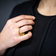Frauenhand mit einem vergoldeten Ring. Dieser besteht aus einer kreisrunden Scheibe, hat eine Öffnung außermittig gesetzt, durch welche der Finger gesteckt ist. Insgesamt ist die Scheibe so aufgewölbt, dass