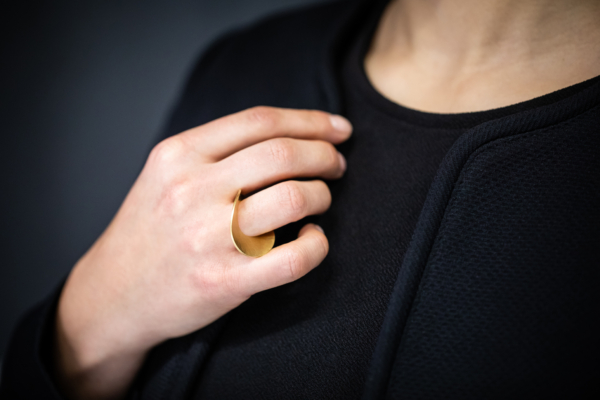 ring-courbe-golden Frauenhand mit einem vergoldeten Ring. Dieser besteht aus einer kreisrunden Scheibe, hat eine Öffnung außermittig gesetzt, durch welche der Finger gesteckt ist. Insgesamt ist die Scheibe an der Mittelachse so aufgewölbt, dass die Ränder nach oben gezogen sind und so die Trageeigenschaft zwischen den Fingern begünstigen.