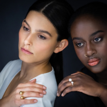 Zwei Frauen mit dem Rücken zueinander und das Gesicht nach vorne gewandt, haben jeweils eine Hand auf ihrer Schulter liegen. An dieser Hand tragen sie einen Zwischenfingerring. Die Ringe sind nach oben hin offen, beschreiben ein U, auf dessen Enden eine Kugel sitzt. Diese liegt dann zwischen den Fingern.