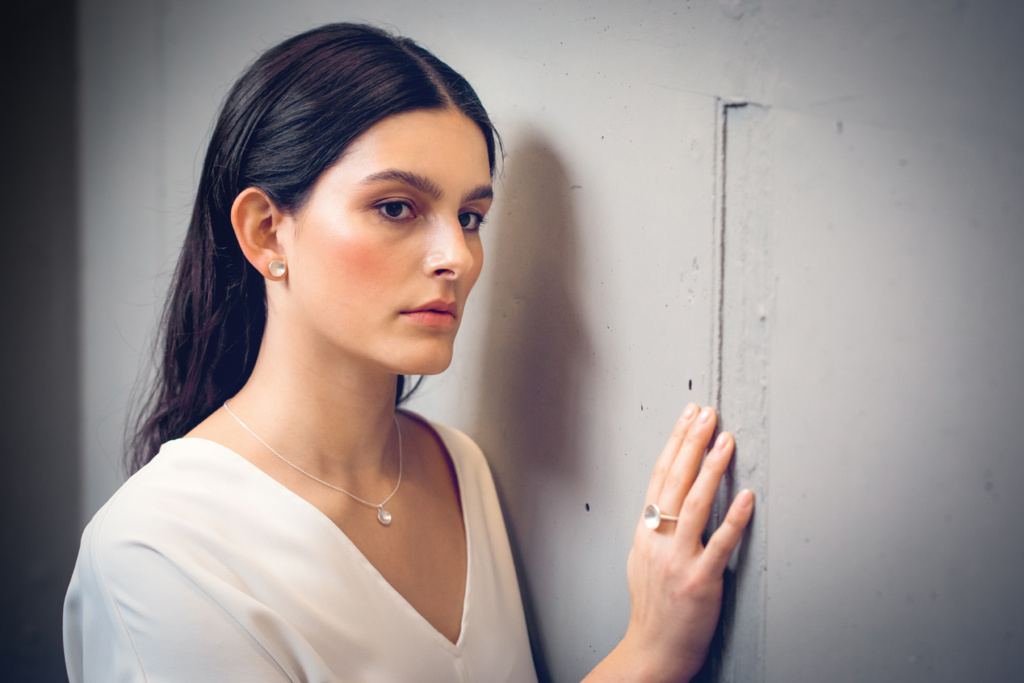 Frau mit langen dunklen Haaren steht an Wand und hat eine Hand an diese gelegt. Sie trägt kleine Silberne Ohrstecker, einen Anhänger an einem zarten Kettchen und einen Ring am Mittelfinger. Alle Schmuckstücke sind aus Silber und in Form einer Schale. 