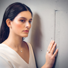 Woman with long dark hair stands on the wall and has put a hand on it. She wears small silver earstuds, a pendant on a delicate chain and a ring on the middle finger. All jewelry is made of silver and in the shape of a shell.