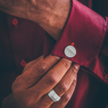 Ring&Manschettenknöpfe_SAZ_72 Männerhand mit breitem Silberring greift die Manschette eines dunkelroten Hemdes an der anderen Hand. Die Manschette ist mit einem zum Ring passenden, kreisförmigen Manschettenknopf geziert.