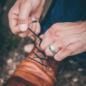 Männerhand bindet Schuhe. Linker Ringfinger ist mit einem auffälligen Silberring geschmückt. Die Oberfläche des breiten Rings ist mit zahlreichen Facetten gestaltet.