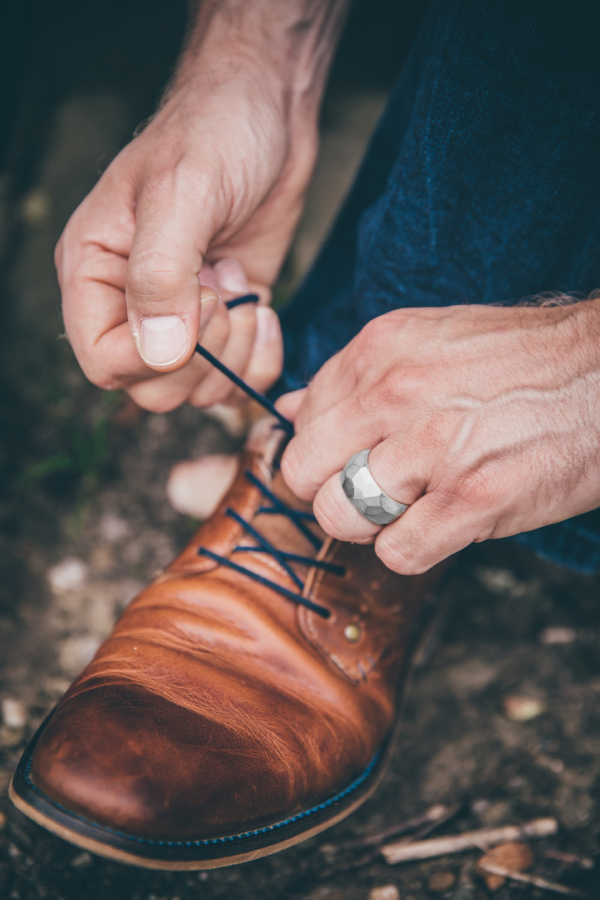 Männerhand bindet Schuhe. Linker Ringfinger ist mit einem auffälligen Silberring geschmückt. Die Oberfläche des breiten Rings ist mit zahlreichen Facetten gestaltet.