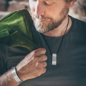 Man with green jacket over his shoulder. The holding hand is adorned with a broad silver bangle, which is embossed with words. Around his neck on a leather strap he wears a silver pendant, also hallmarked with letters.