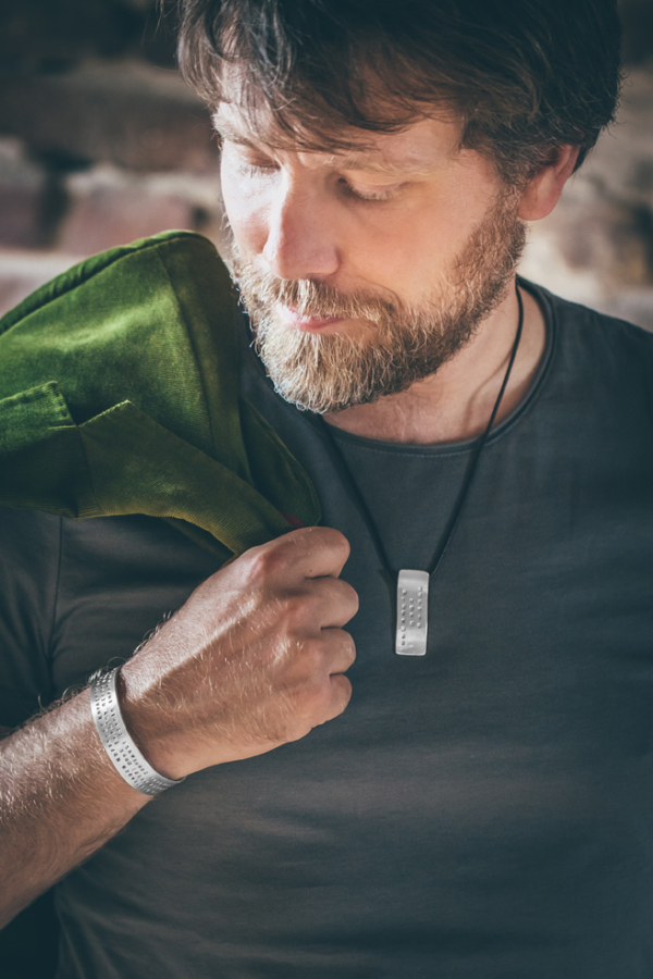 armreif&anhaenger_text_silber Mann mit grünem Jacket über der Schulter. Die haltende Hand ist mit einem breiten Silberarmreif geschmückt, welcher mit Wörtern geprägt ist. Um den Hals an einem Lederband trägt er einen Silberanhänger, ebenfalls mit Buchstaben punziert.