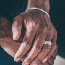 Männerhände aufeinander gelegt. Die obere Hand trägt einen breiten, geschmiedeten Silberring am Ringfinger und am Handgelenk eine schmale und ebenfalls geschmiedete Silberarmspange