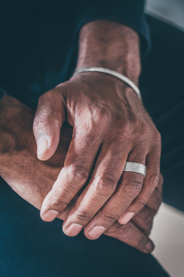 Männerhände aufeinander gelegt. Die obere Hand trägt einen breiten, geschmiedeten Silberring am Ringfinger und am Handgelenk eine schmale und ebenfalls geschmiedete Silberarmspange