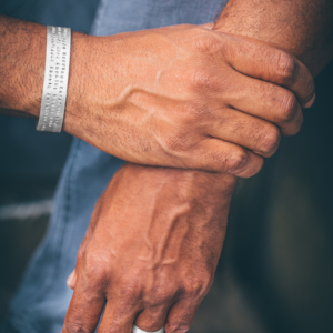 Eine Männerhand umschließt das Handgelenk einer anderen Hand. Am sichtbaren Handgelenk ist ein breiter Silberarmreif in welchen drei Zeilen Text einpunziert sind.