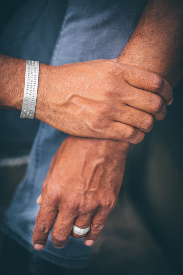 Armreif_text_Ring_SAZ_closeup_72 Eine Männerhand umschließt das Handgelenk einer anderen Hand. Am sichtbaren Handgelenk ist ein breiter Silberarmreif in welchen drei Zeilen Text einpunziert sind.