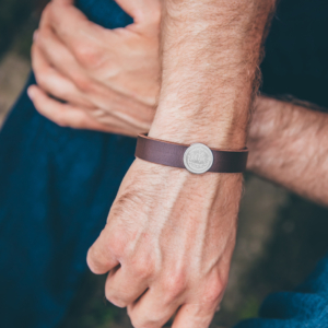 Zwei Männerhände über Kreuz. Die vordere Hand hat um das Handgelenk ein braunes Lederarmband mit verziertem Schiebebutton aus Silber.