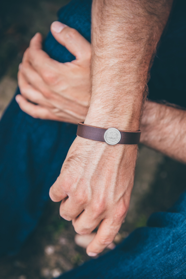 Lederarmband_Zierbutton_72 Zwei Männerhände über Kreuz. Die vordere Hand hat um das Handgelenk ein braunes Lederarmband mit verziertem Schiebebutton aus Silber.