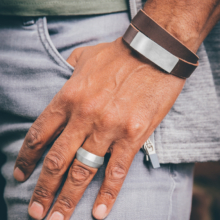 Männerhand mit breitem Silberring und doppelt gewickeltem Armband aus braunem Leder. Eine rechteckige Schließe, so breit wie das Armband und etwa 50mm lang ist Zier- und Funktionselement zugleich.