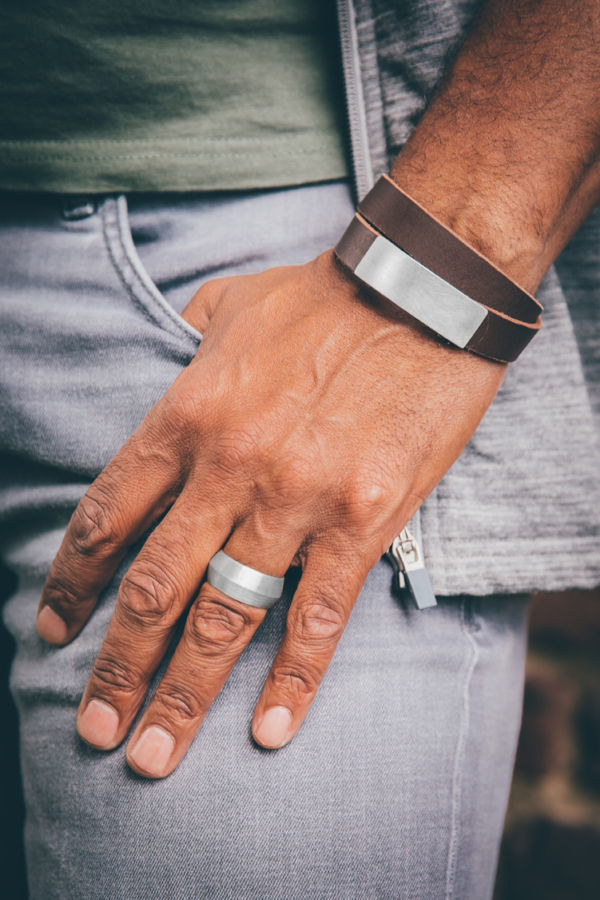 Man's hand with a wide silver ring and a double-wrapped bracelet of brown leather. A rectangular clasp, as wide as the bracelet and about 50mm long, is both a decorative and functional element.
