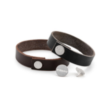 lederarmbänder-textbutton-silber Zwei Lederarmbänder mit Silberbuttons. Diese sind mit Wörtern geprägt. Zwei weitere Silberbuttons liegen davor. Einer ebenfalls beschriftet, der andere ist von der Rückseite gezeigt.
