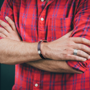 Verschränkte Männerellenbogen. Die sichtbare Hand ist mit einem Silberring und am Handgelenk mit einem Lederarmband geschmückt. Das Lederarmband ist mit einem Silberbutton versehen, welcher mit Buchstaben punziert ist. Auch der Silberring ist mit Wörtern punziert