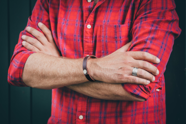 Crossed male elbows. The visible hand is adorned with a silver ring and at the wrist with a leather bracelet. The leather bracelet has a silver button which is punched with letters. The silver ring is also hallmarked with words