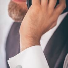 Männerhand mit Handy am Ohr. Das Handgelenk zeigt ein weißes Manschettenhemd, geziert von silbernen Manschettenknöpfen. Diese sind in Form Kreisrunder Scheiben mit einem räumlicher Wirkung durch einen aufgebogenen Spalt.