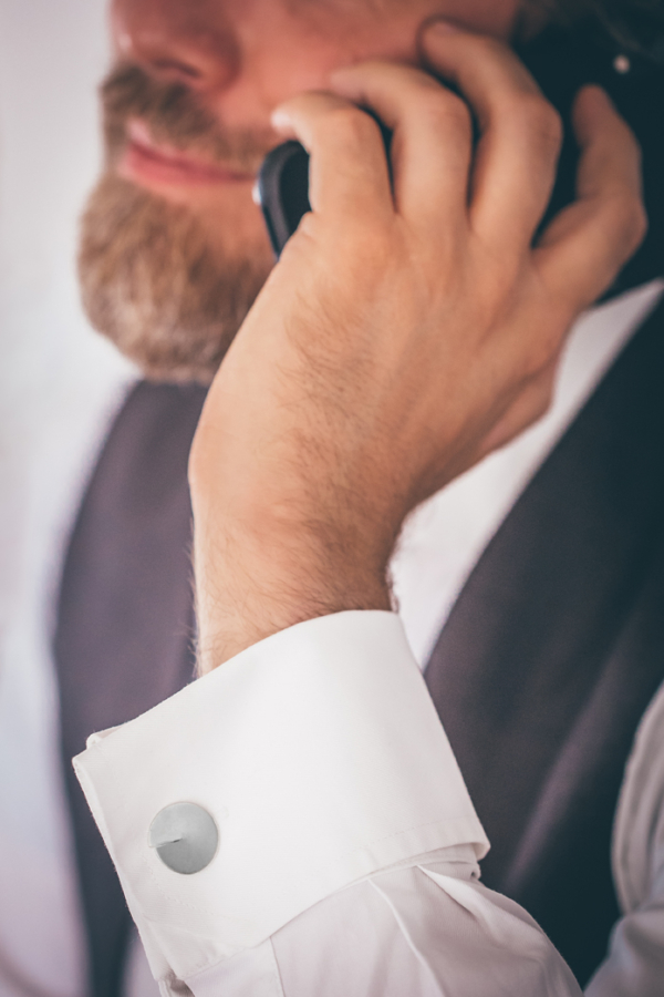 Männerhand mit Handy am Ohr. Das Handgelenk zeigt ein weißes Manschettenhemd, geziert von silbernen Manschettenknöpfen. Diese sind in Form Kreisrunder Scheiben mit einem räumlicher Wirkung durch einen aufgebogenen Spalt.