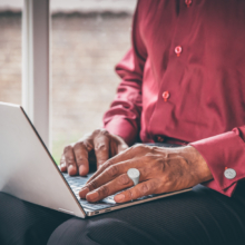 Ring&Manschettenknöpfe_Siegel_Laptop_72 Männerhände auf einer Computertastatur. Die vordere Hand ist von einem silbernen Siegelring und an der Manschette des dunkelroten Hemdes mit einem Manschettenknopf geziert. Beide Schmuckstücke sind mit einem Wappen graviert.