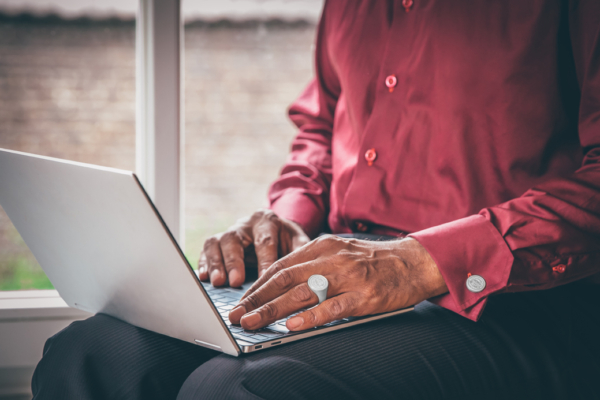 Ring&Manschettenknöpfe_Siegel_Laptop_72 Männerhände auf einer Computertastatur. Die vordere Hand ist von einem silbernen Siegelring und an der Manschette des dunkelroten Hemdes mit einem Manschettenknopf geziert. Beide Schmuckstücke sind mit einem Wappen graviert.