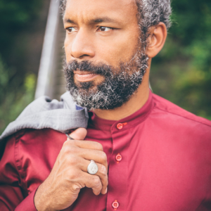 Mann mit dunkelrotem Hemd und über der Schulter ein graues Jacket. Die sichtbare Hand ist am Ringfinger von einem silbernen Siegelring geschmückt.