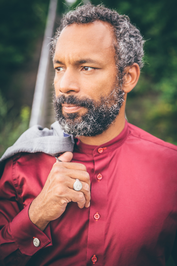 Siegelring-Silber Mann mit dunkelrotem Hemd und über der Schulter ein graues Jacket. Die sichtbare Hand ist am Ringfinger von einem silbernen Siegelring geschmückt.