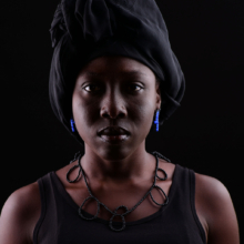 kette_capreolo_ohrst_tendrilblau Frau mit Kette und Ohrschmuck aus kleinen Onyxperlen. Diese sind so geknüpft, dass unterschiedlich große Zier-Schlaufen entlang der Kette entstehen. Der Ohrschmuck ist zusätzlich aus blauen Achatperlen. Die Schwarzen und blaue Perlen sind zu zwei Schlaufen auf Nylon aufgezogen und mit einem Stecker aus Silber versehen.