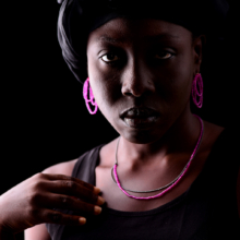 ohrschmuck+kette_rosea Porträt einer Frau, die pinken Ohrschmuck und eine schwarz pinke Kette trägt. Die Kette ist zweireihig: einmal Rubelit und einmal geschwärzte Silberkügelchen, der Ohrschmuck aus drei untereinanderhängenden Schlaufen aus Rubelit auf Nylon und ist mit einem Silberstecker versehen.